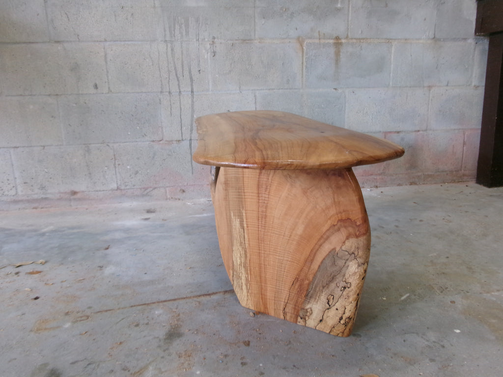 Wooden stool with a natural, curved design against a stone wall.